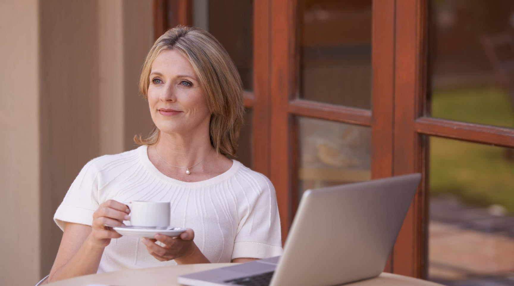 Middle-aged woman sitting outdoors with a laptop and a cup of coffee, looking thoughtfully to the side