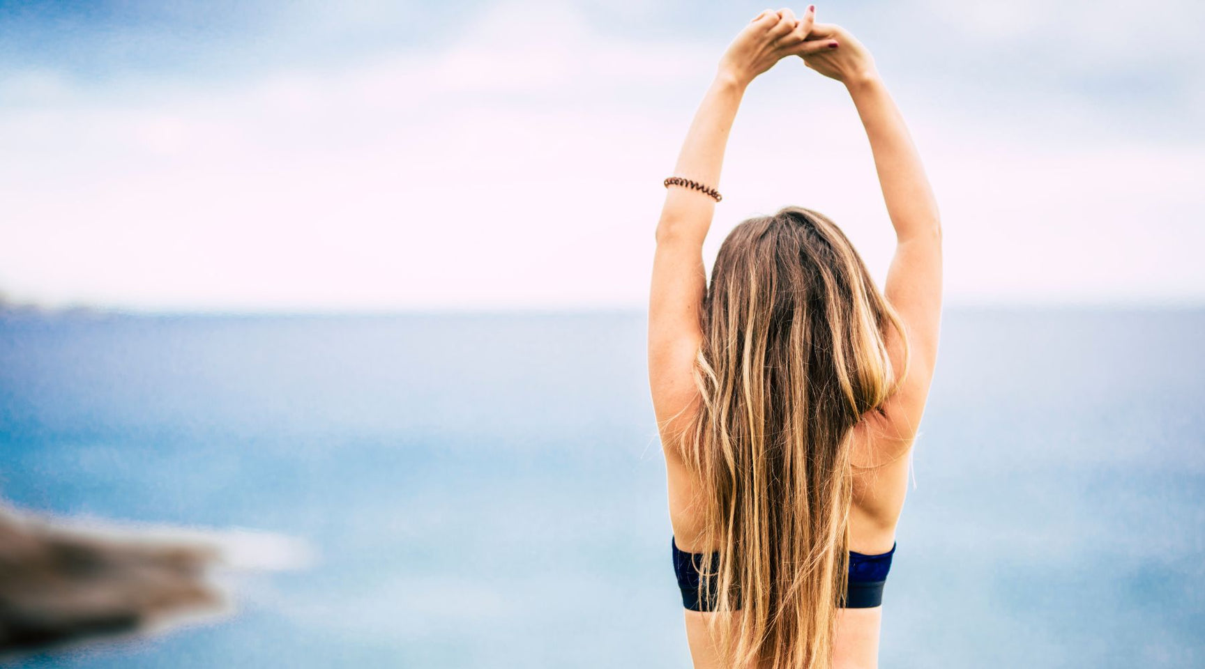 Woman with long hair stretching with arms raised while standing near an ocean coastline
